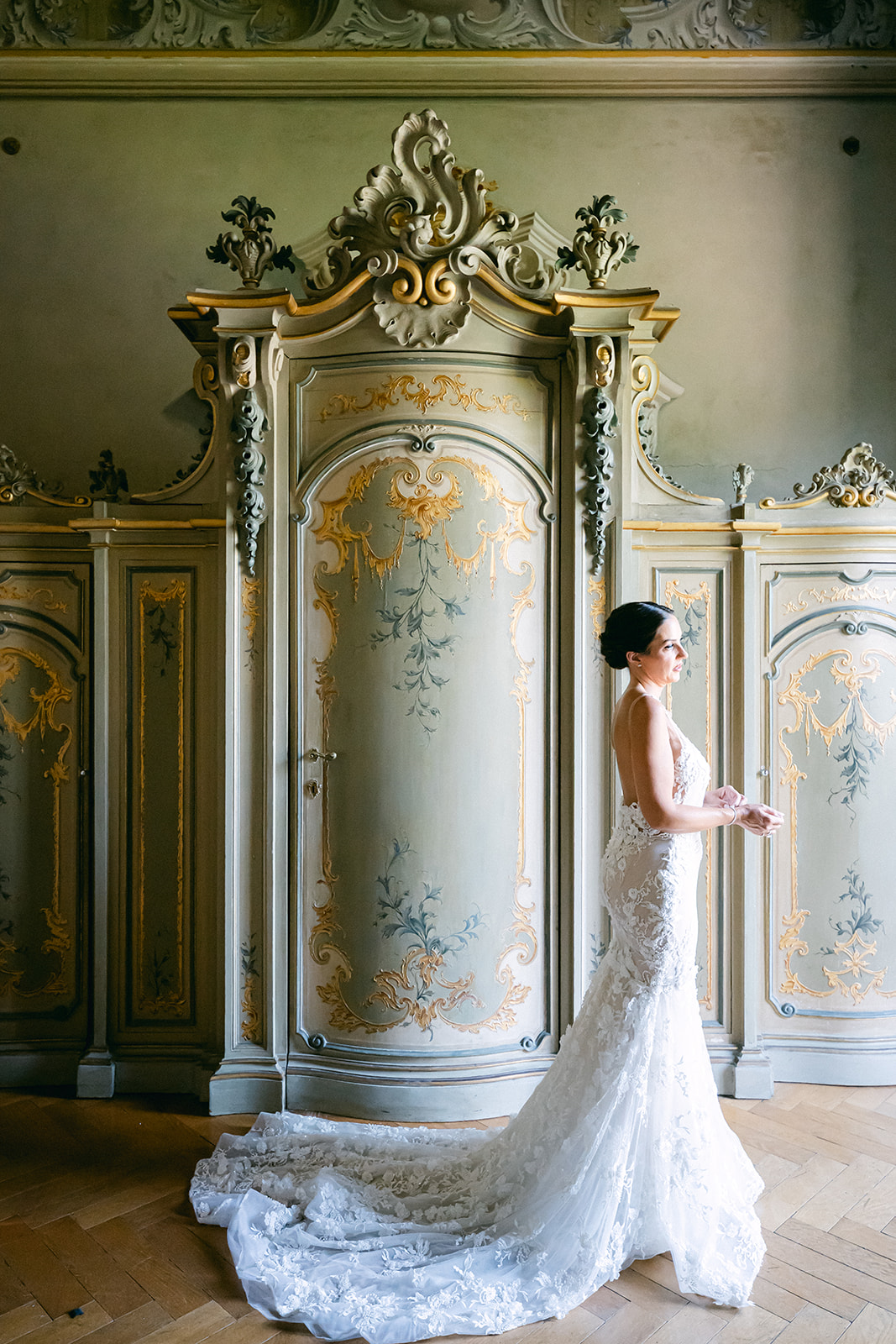 Fran in her lace wedding gown standing beside an ornate painted cabinet at Villa Erba.