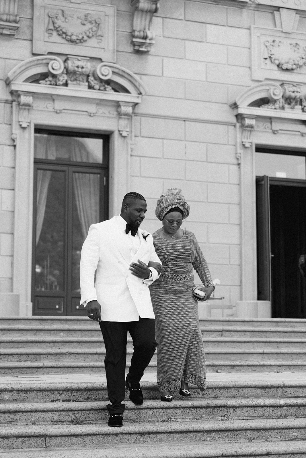Black and white photograph of Ope walking down Villa Erba's steps beside a woman in a gold dress.