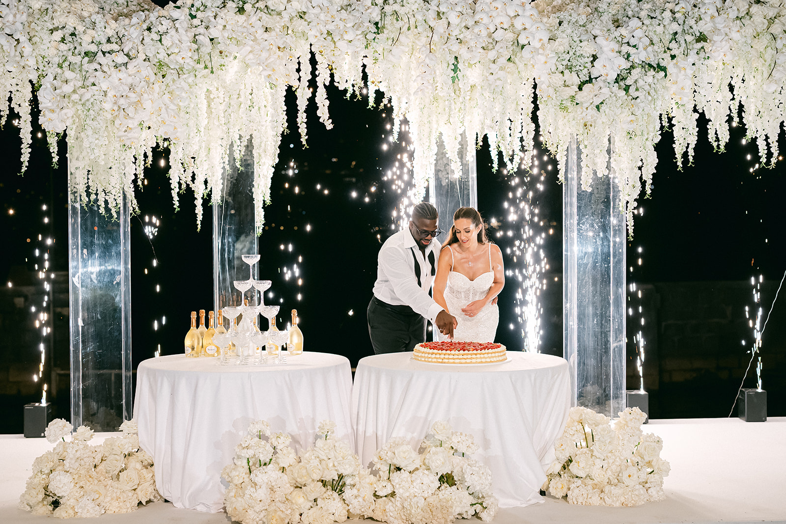 Fran and Ope celebrating beside their wedding cake beneath cascading white flowers at night.